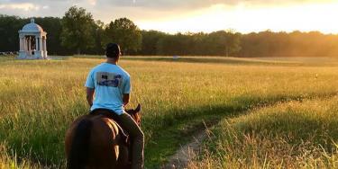 A tall grassy field with the sun on the horizon with a horseback rider ahead of another. 