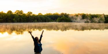 A hiker with her arms raised overlooking a lake at sunset